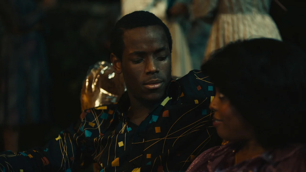 Lover’s Rock (2020) movie still: medium — A dimly lit indoor space with blurred figures in the background. A man sits while looking at the woman…