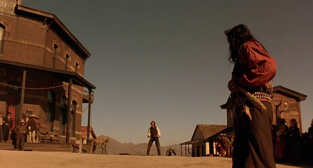 The Quick and the Dead (1995) movie still: wide — A dusty, 19th-century frontier town street is flanked by wooden and brick buildings. Two men stand…