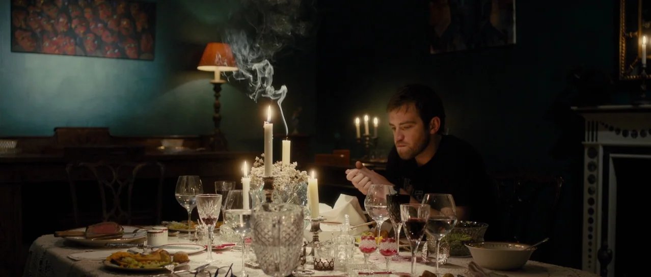 Road Games (2015) movie still: medium — A dark dining room features a long table set for a formal meal. A man sits alone at the head of the table…