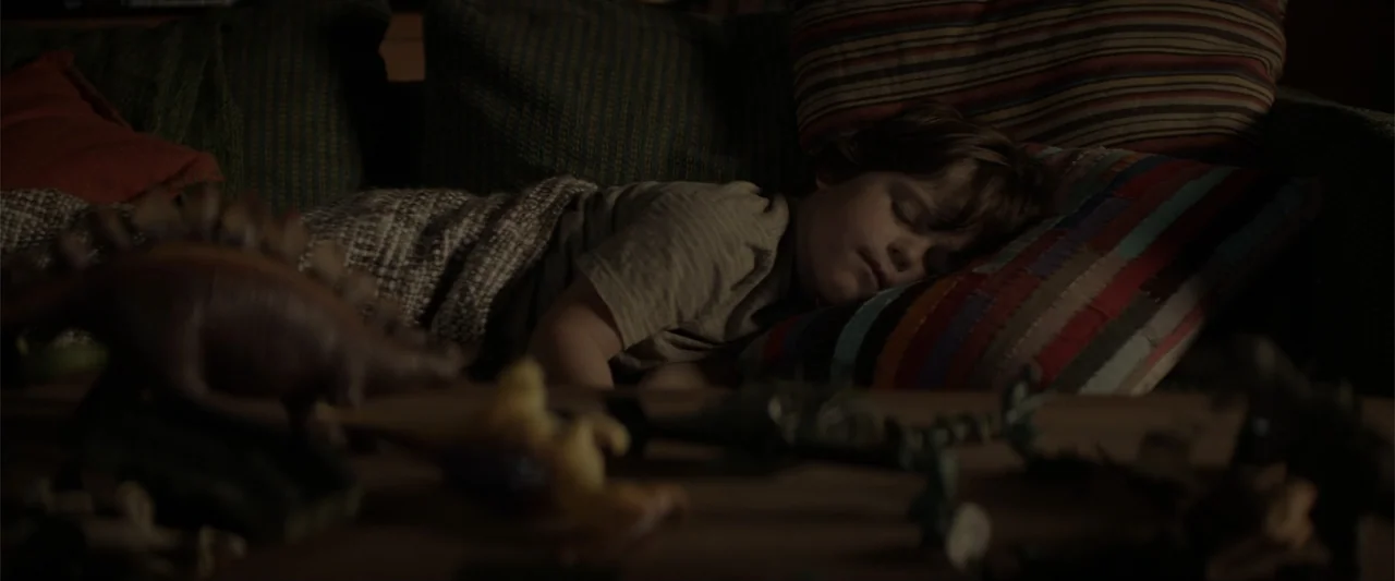 Godzilla (2014) movie still: medium — A dark living room features a child asleep on a couch draped in a blanket. Various toy dinosaurs are scattered…