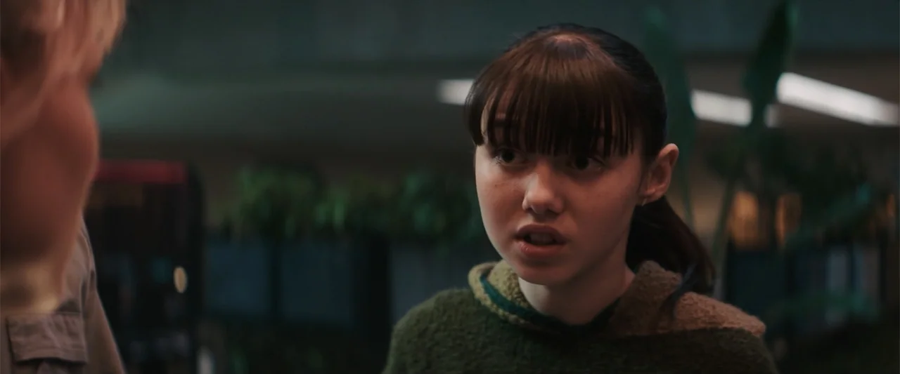 M3GAN 2.0 (2025) movie still: closeup — An interior office space with blurred green foliage in the background. A young woman with blunt bangs looks…