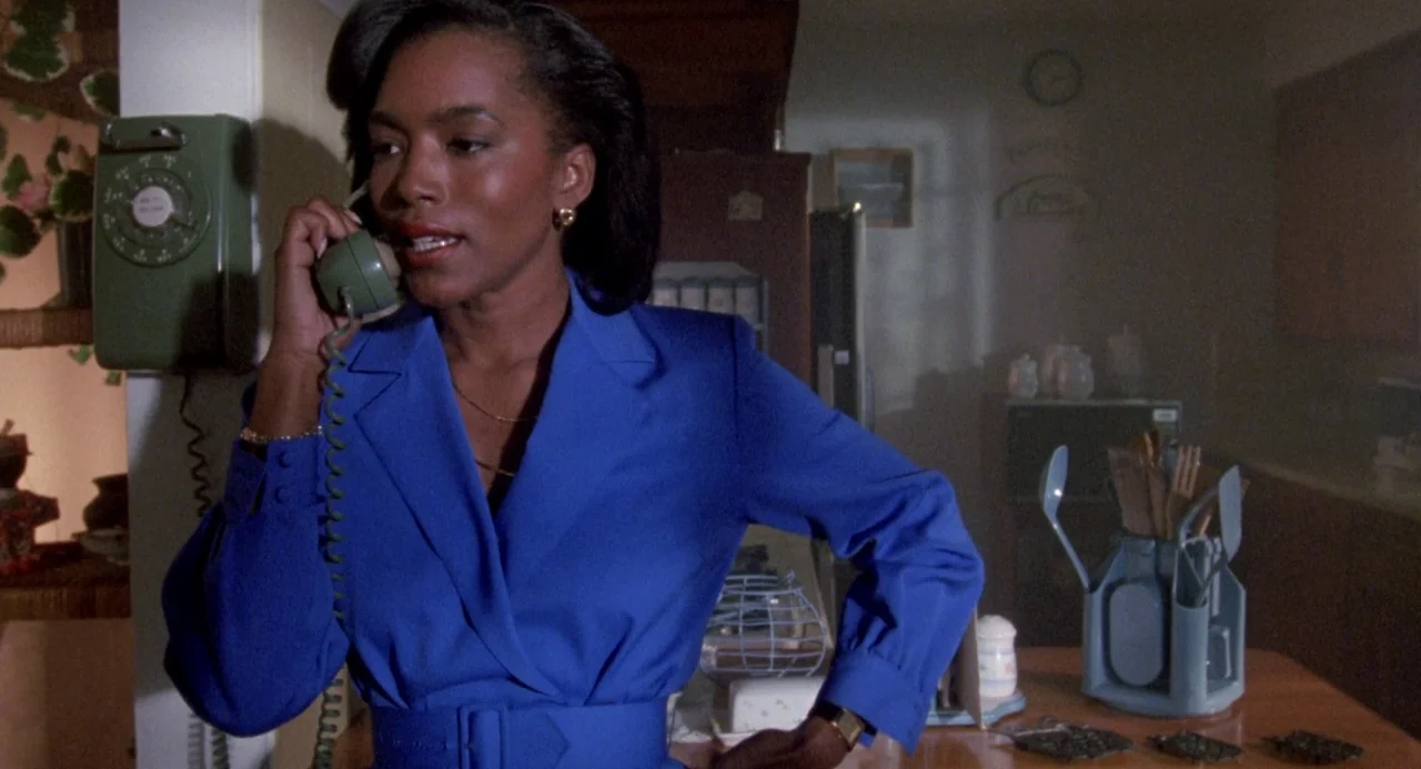 Boyz In The Hood (1991) movie still: medium — A dimly lit room with wooden cabinets and a wall-mounted rotary telephone. A woman in a blue blazer…