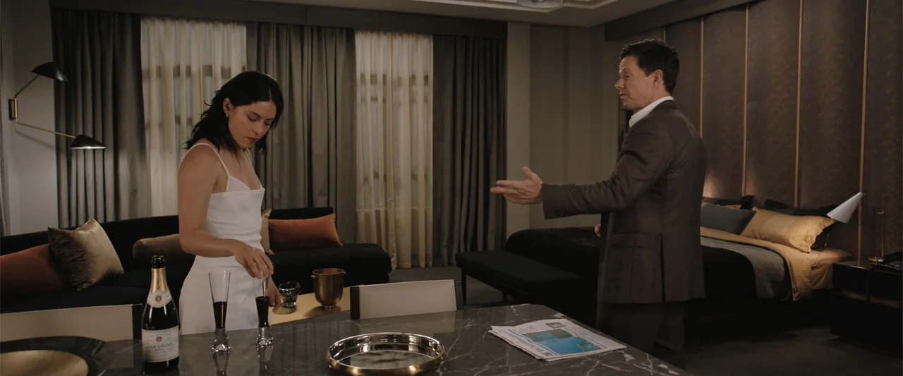 Play Dirty (2025) movie still: medium — A luxury hotel suite contains a marble table with champagne and a bed background. A man in a brown suit…