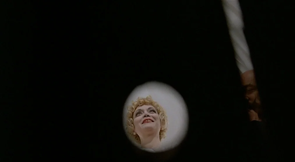 The Voice of the Moon (1990) movie still: closeup — A woman's face is seen through a small round peephole in a dark door. The subject smiles upward…