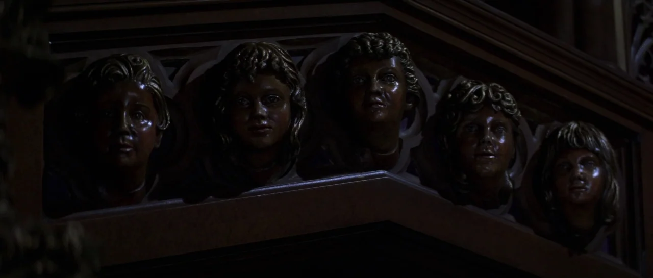 The Haunting (1999) movie still: closeup — Five identical carved wooden heads of children are embedded into a decorative structural beam. The faces…