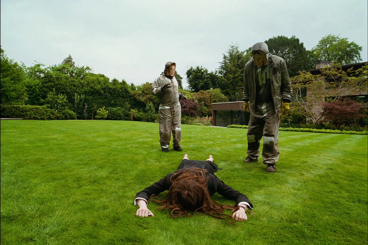 Bugonia (2025) movie still: wide — A manicured green lawn extends toward a modern house in the background. Two masked figures wearing industrial…