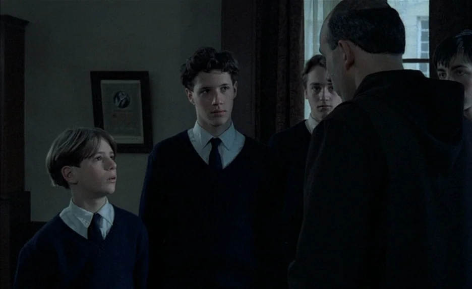 Au Revoir Les Enfants (1987) movie still: medium — A dimly lit interior space with students standing before a teacher in black robes. The…