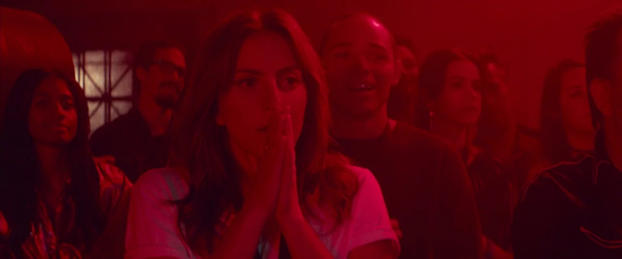 A Star Is Born (2018) movie still: medium — A crowded venue interior is saturated in intense red light. A woman holds her hands together in front of…