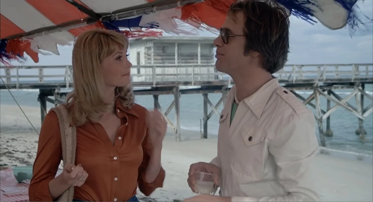 Empire of the Ants (1977) movie still: medium — A wooden pier extends into the ocean behind a covered picnic area on sand. A blonde woman and a man…