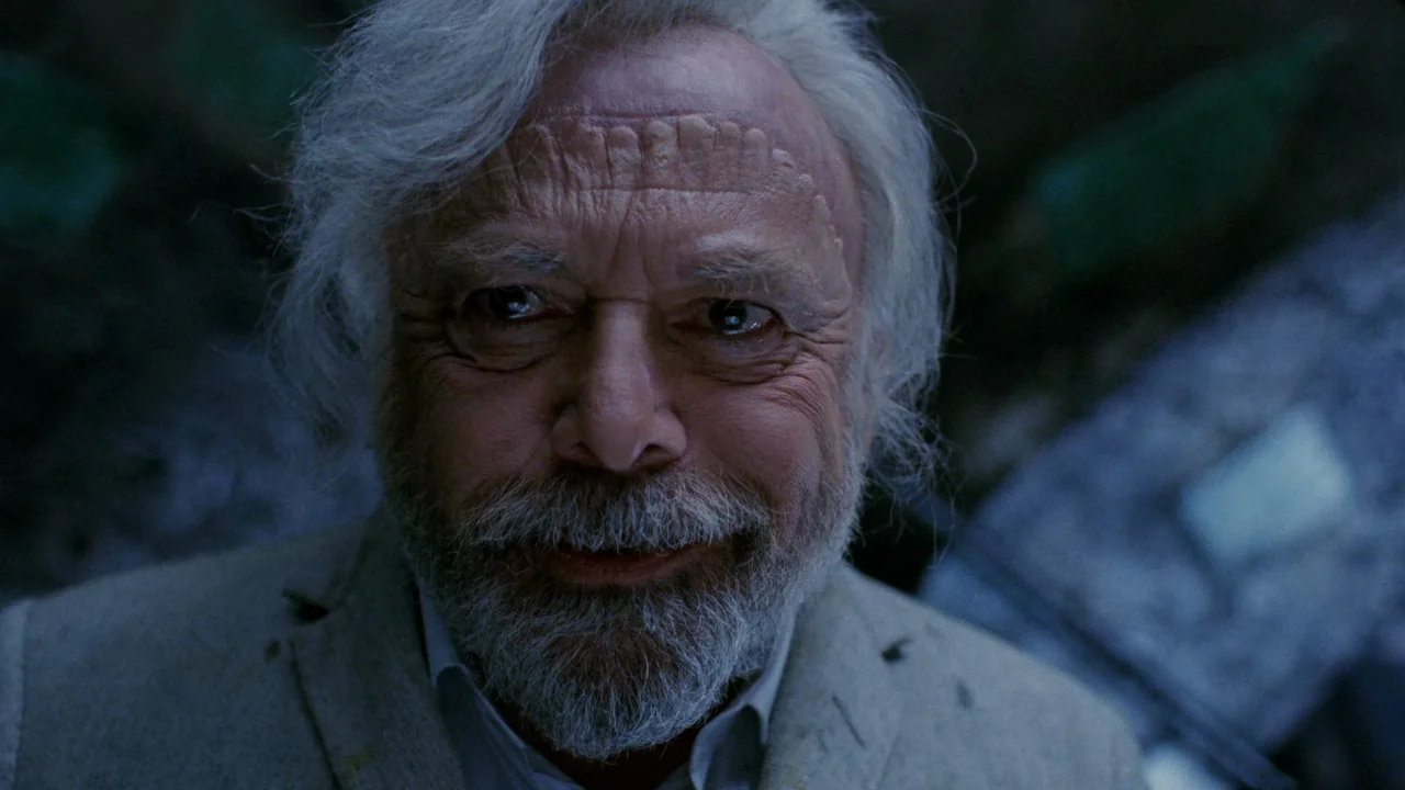 The Sect (1991) movie still: closeup — An elderly man is framed closely against a dark, out-of-focus dense green background. He looks forward with a…