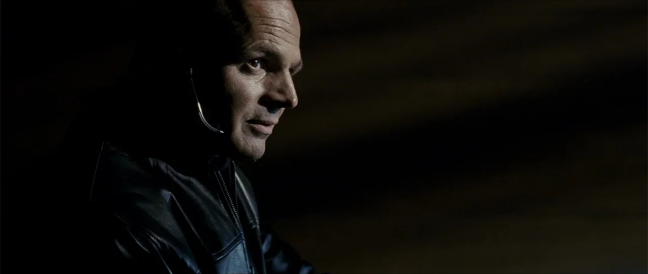 Elektra (2005) movie still: closeup — A man stands in an underexposed environment with dark surroundings. The subject wears a headset and a black…