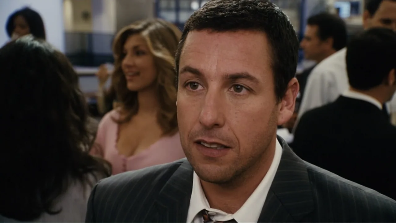 Click (2006) movie still: medium — A man in a suit stands in a brightly lit room filled with other people. He is the central subject as he speaks to…
