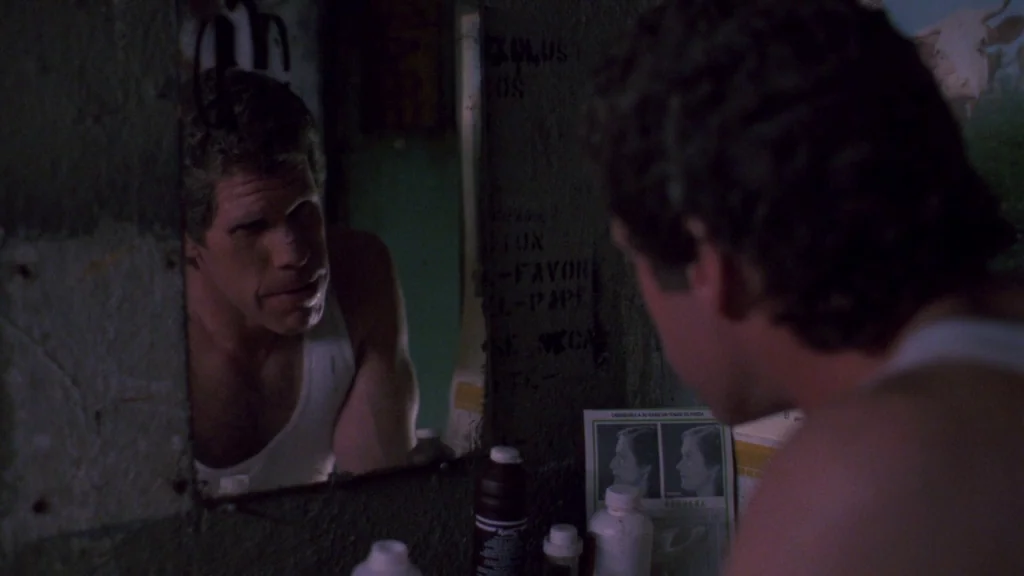 Cronos (1993) movie still: medium — A dimly lit room features a small mirror mounted on a textured, dark wall. A man in a white tank top gazes into…