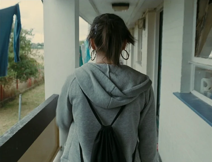 Fish Tank (2009) movie still: medium — An exterior concrete building walkway with laundry hanging on a line in the background. A young woman in a…