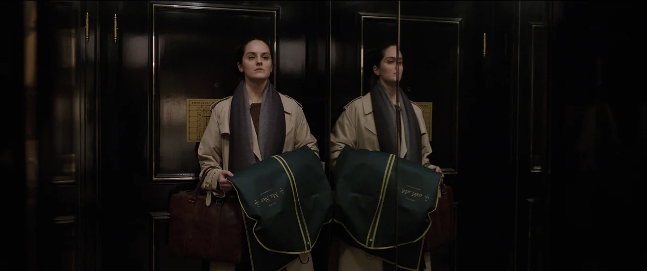 Tár (2022) movie still: medium — A woman stands in a high-end wood-paneled elevator with a brass trim detail. She holds a green garment bag and a…