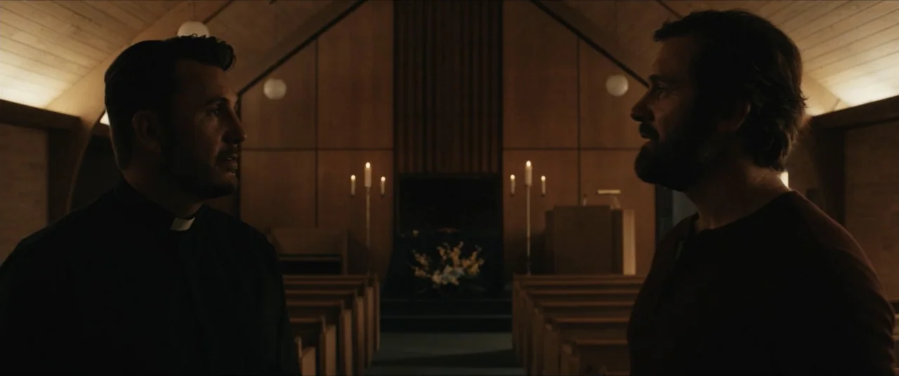 Possessions (2024) movie still: medium — A dimly lit chapel interior with wooden pews facing a central altar area. Two men stand in the foreground…