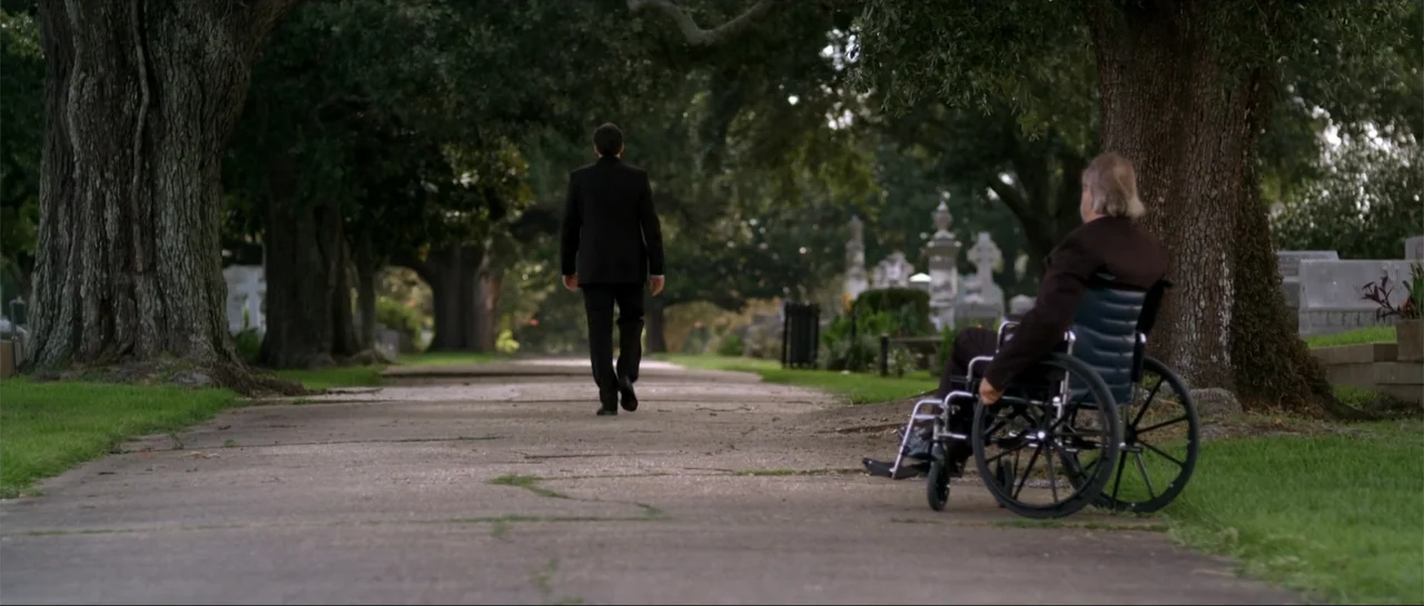 Rage (2014) movie still: wide — An outdoor cemetery path is flanked by large trees and aged stone gravestones. A man in a suit walks away while a…