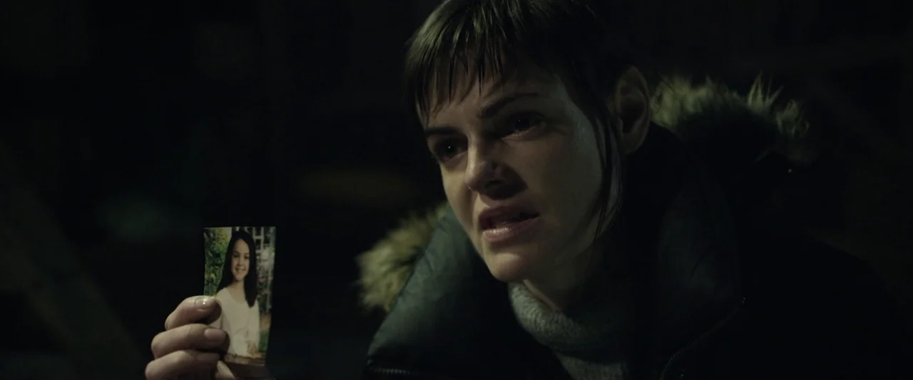 Motherly (2022) movie still: closeup — A darkened interior space provides little environmental detail beyond shadow. A woman in a dark winter coat…