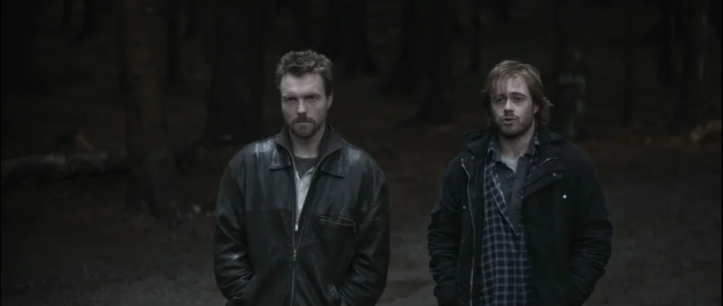 One Hundred Mornings (2009) movie still: medium — Two men stand in a dense, dark forest with muted colors and trees. They look directly forward…