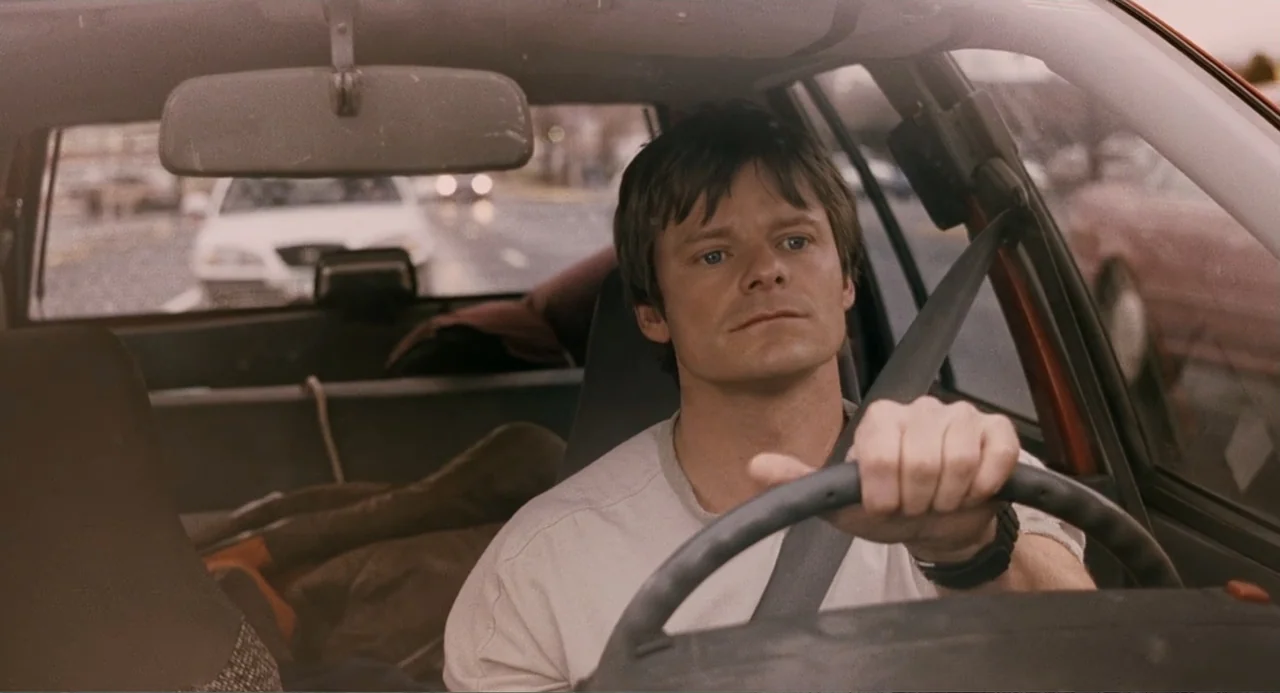 Management (2009) movie still: medium — A man driving a car during daylight hours while viewed from the passenger seat. The interior of the vehicle…