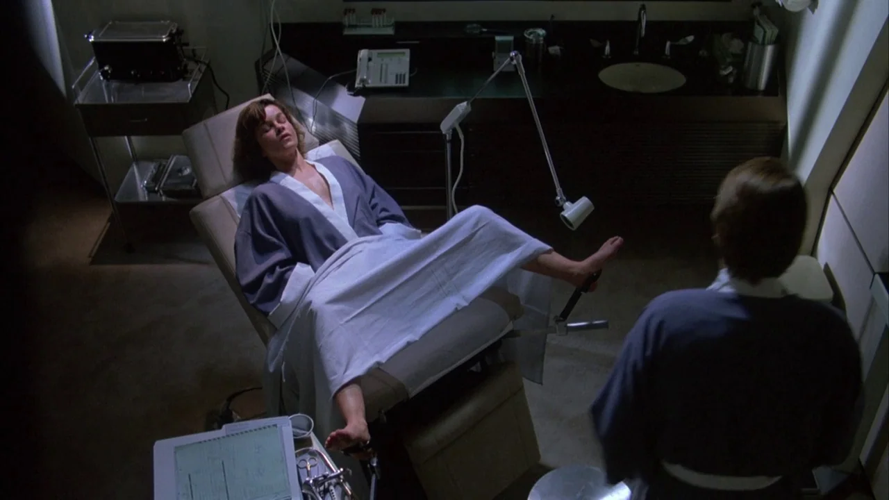 Dead Ringers (1988) movie still: medium — A medical exam room features dark cabinetry and clinical equipment on clean counters. A woman rests on the…
