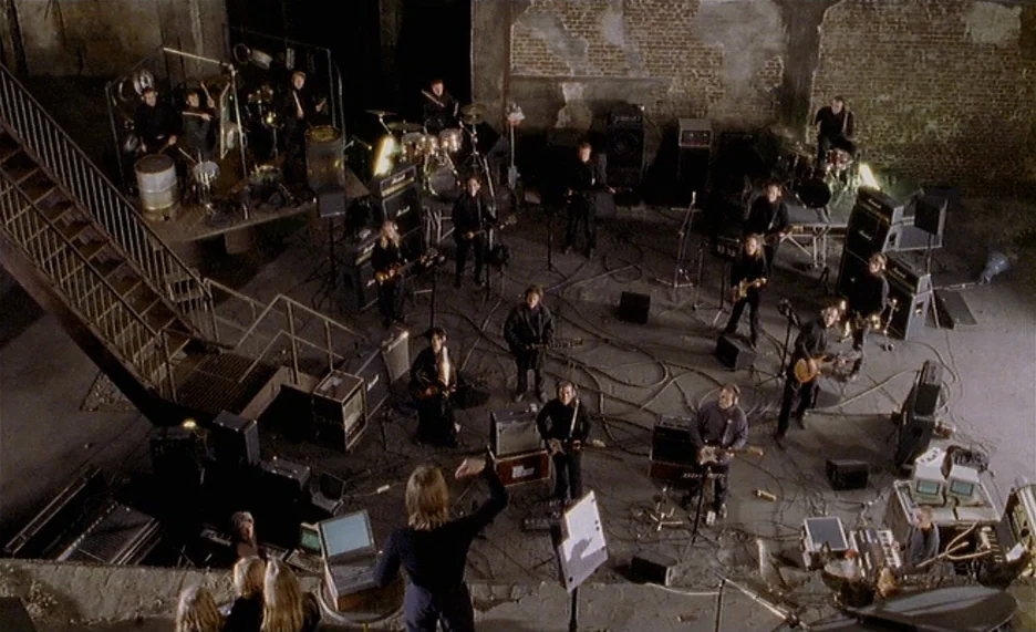 Pola X (1999) movie still: wide — A wide industrial space features exposed brick walls and a metal staircase platform. Numerous musicians surround…