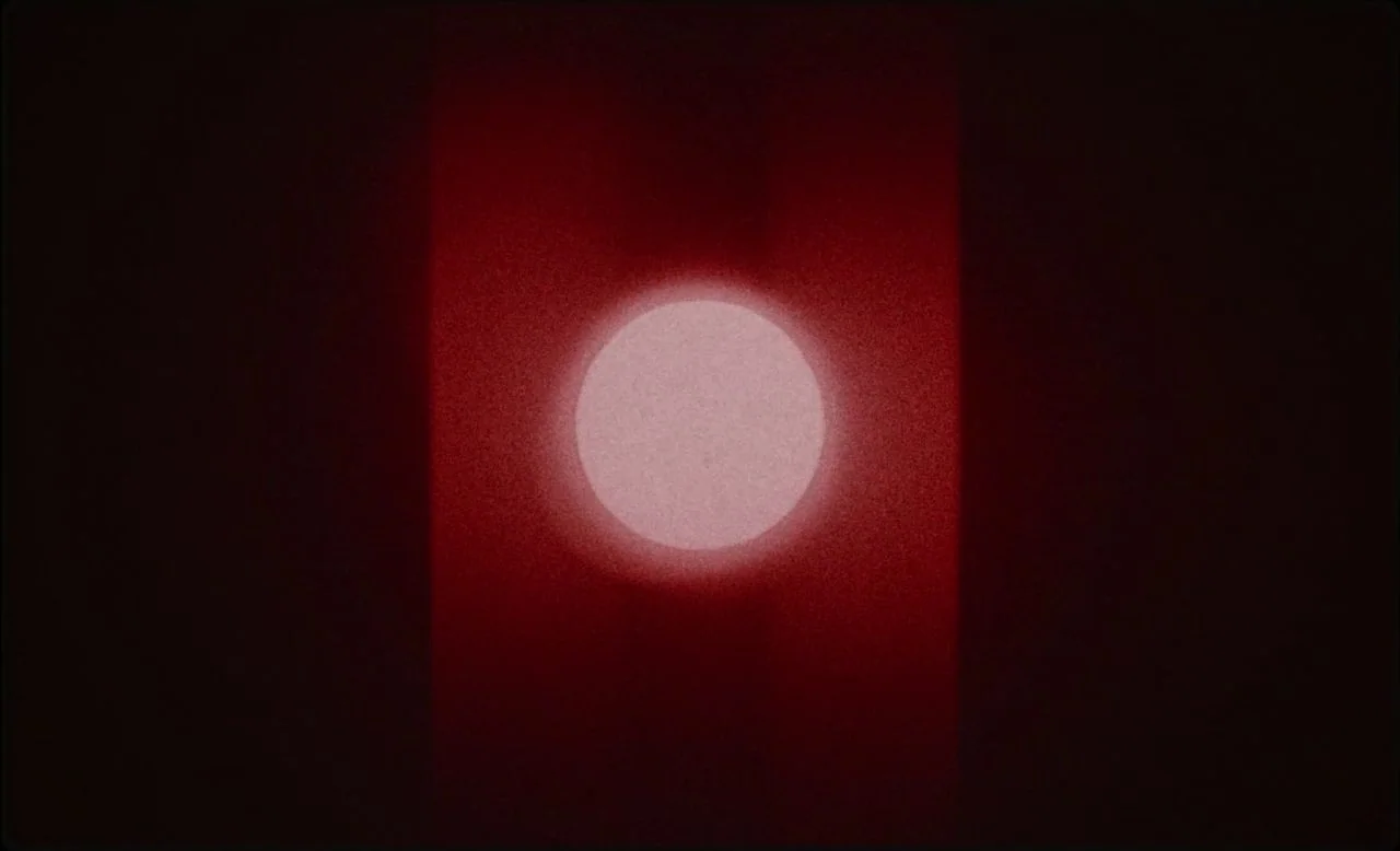 Last and First Men (2020) movie still: establishing — A dark, textured void is bisected by a vertical band of deep crimson light. A single bright…
