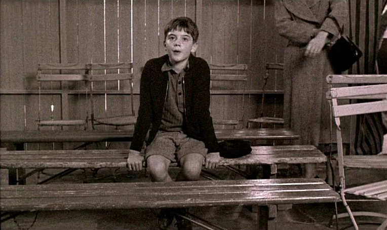 Jacquot de Nantes (1991) movie still: medium — A sparsely furnished waiting area features rows of wooden benches against a vertical wall. A young…