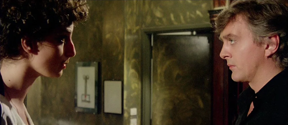 Deep Red (1975) movie still: medium — A dimly lit hallway features walls with mottled green paint and framed pictures. Two people stand facing each…