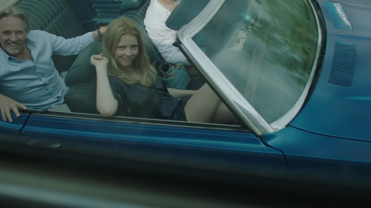 Infinity Pool (2023) movie still: medium — A deep blue convertible car cruises outdoors during the daytime. A blonde woman and an older bearded man…