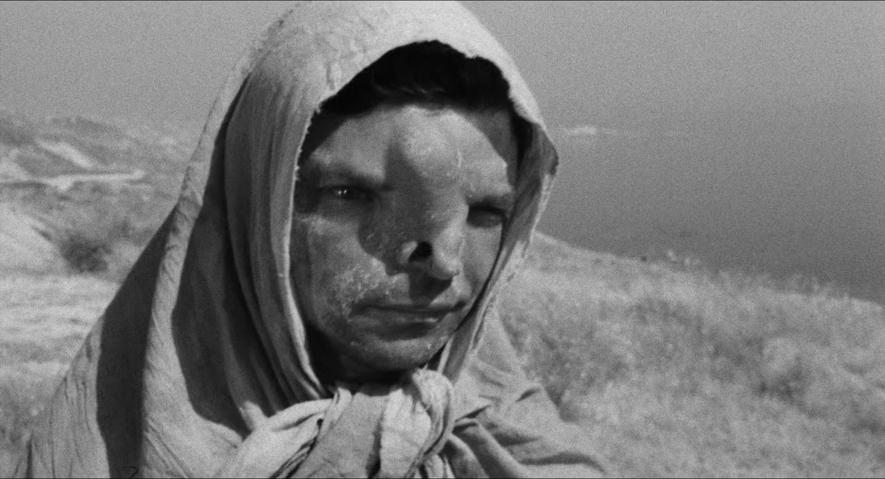 The Gospel According To St. Matthew (1964) movie still: closeup — A desolate, dry hillside with sparse vegetation under a bright, cloudless sky. A…