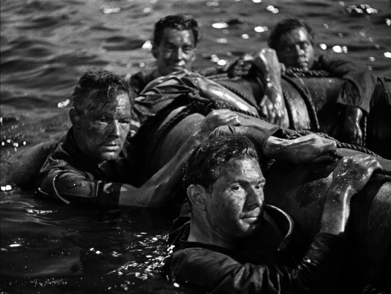 In Which We Serve (1942) movie still: medium — A group of men struggle in dark ocean water while clinging to a life raft. Sunlight glints off their…