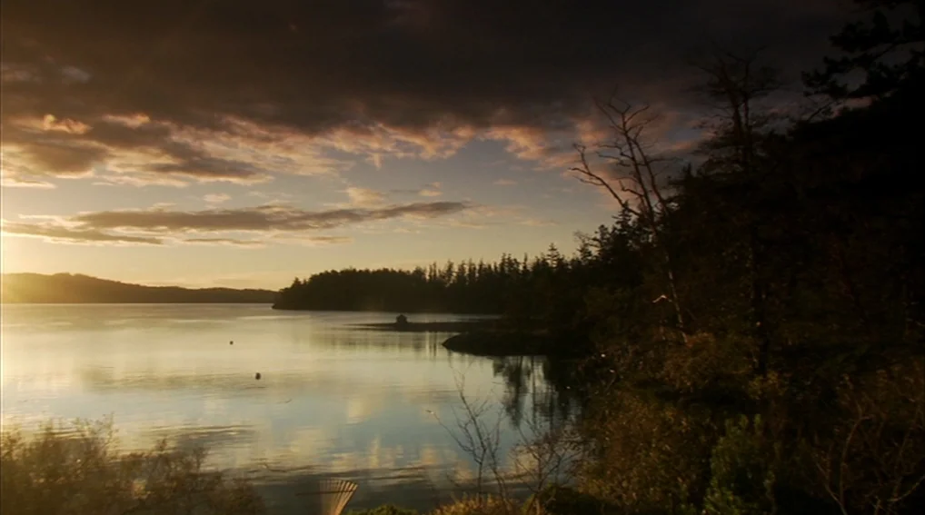Your Sister’s Sister (2011) movie still: establishing — A calm lake stretches toward distant hills under a sunset sky with scattered clouds. A dark,…