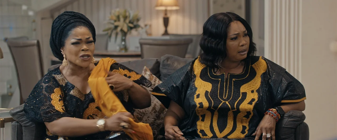 Big Love (2012) movie still: medium — A well-lit living room features upholstered furniture and a table lamp in background. Two middle-aged women…