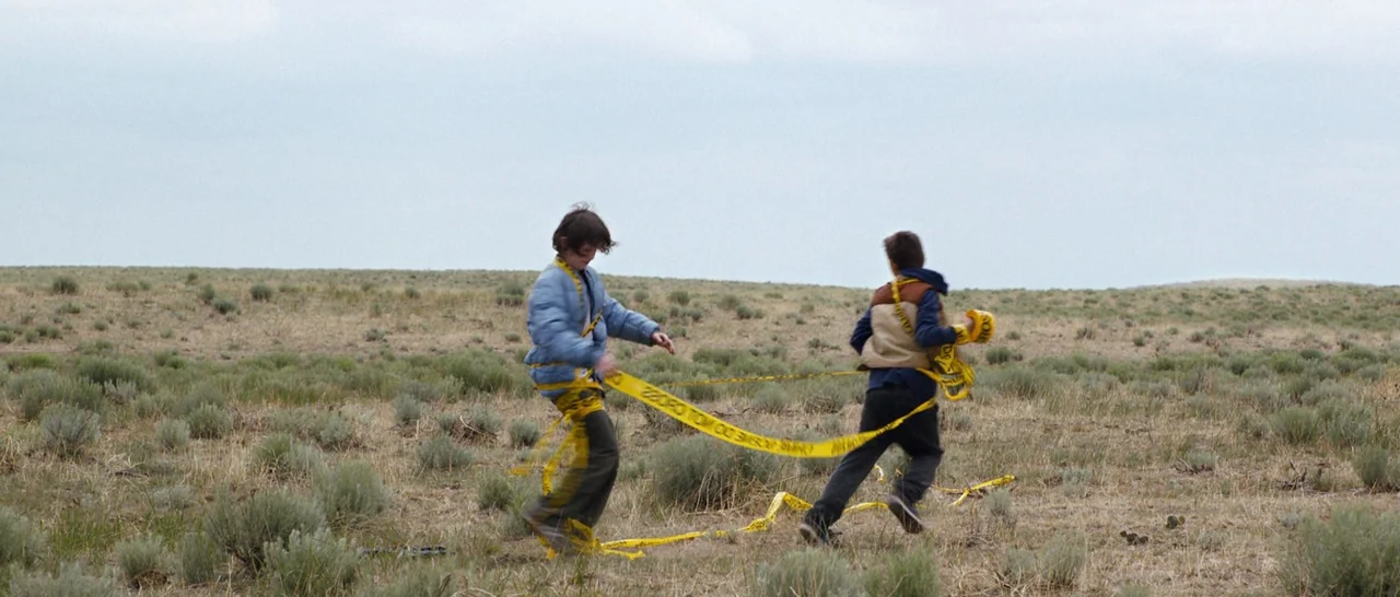 Cop Car (2015) movie still: wide — A vast, open plain covered in sparse sagebrush and dry, yellowing grasses. Two boys run through the landscape…