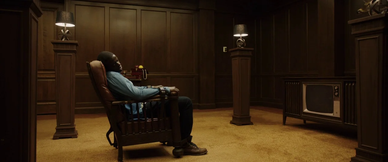 Get Out (2017) movie still: wide — A dark wood-paneled room is furnished with two pedestal lamps and a vintage television set. A man in a blue shirt…