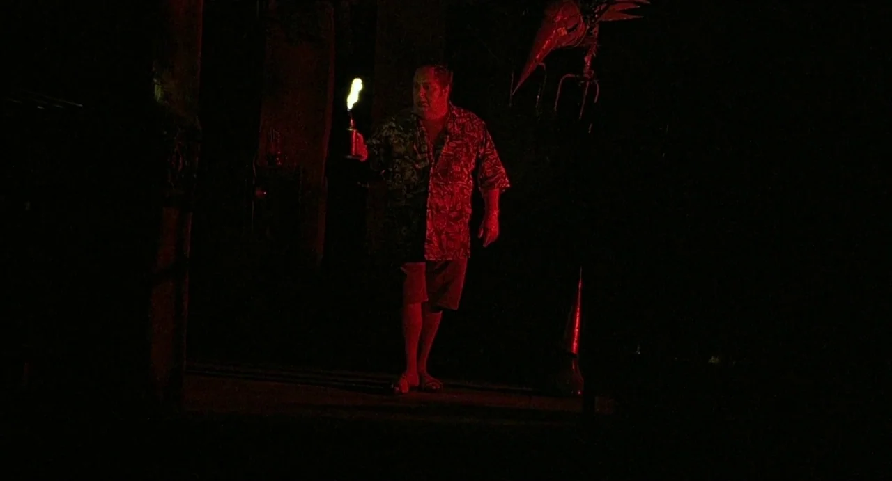Hardware (1990) movie still: fullbody — A dark outdoor space at night features an unidentifiable metal object hanging from above. A man walks…