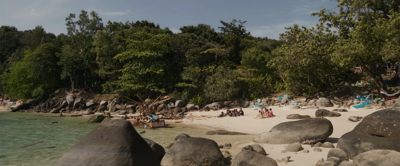 Meg 2: The Trench (2023) movie still: wide — A tropical beach features white sand, large granite boulders, and dense green jungle foliage. A small…