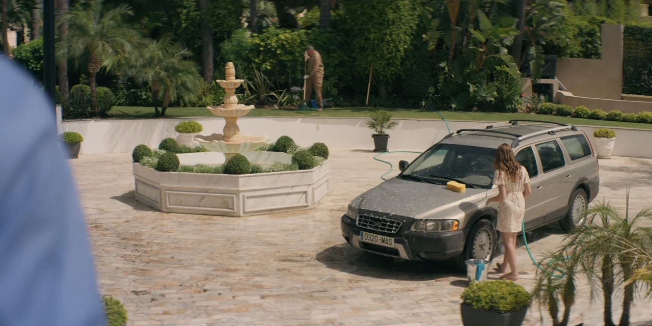 My Fault (2023) movie still: wide — A stone-paved driveway surrounds a tiered marble fountain in a lush estate garden. A woman cleans a station…
