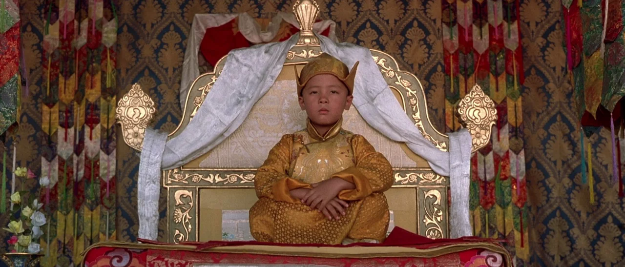 Kundun (1997) movie still: medium — A young boy sits on a gilded throne draped with white silk inside a room. The background features blue patterned…