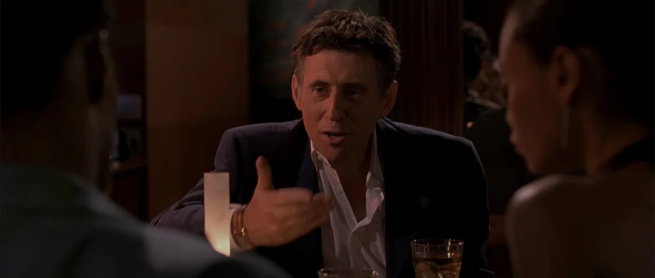 Shade (2003) movie still: medium — A dimly lit restaurant interior with dark walls and blurry background details. Three people are seated at a…