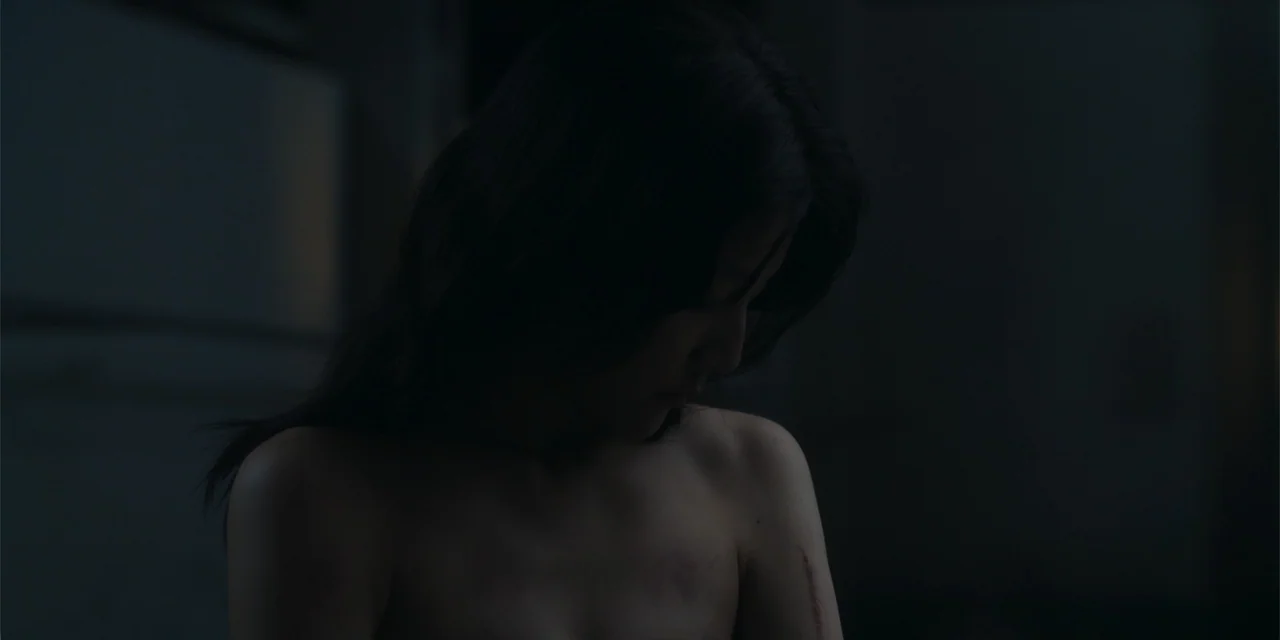 Host (2020) movie still: medium — A dimly lit room with dark walls surrounds the subject in low light. A woman with dark hair looks down at a…
