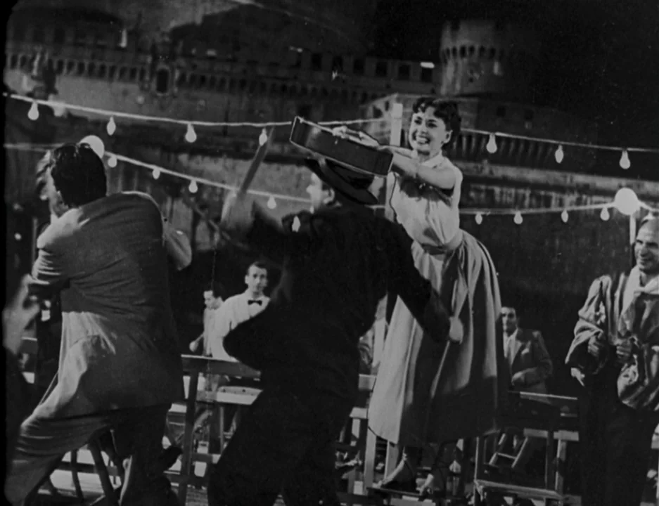Roman Holiday (1953) movie still: medium — An outdoor terrace at night features people dancing under hanging string lights. In the background looms…
