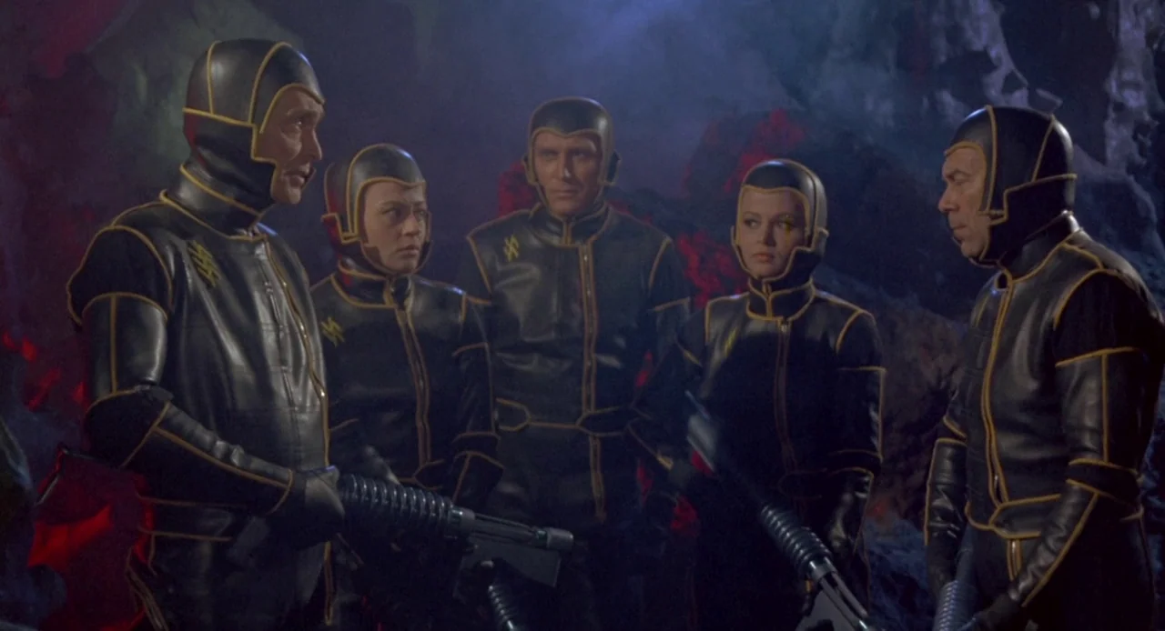 Planet of the Vampires (1965) movie still: medium — A group of five figures stand in a dark, jagged rocky environment at night. They wear black…