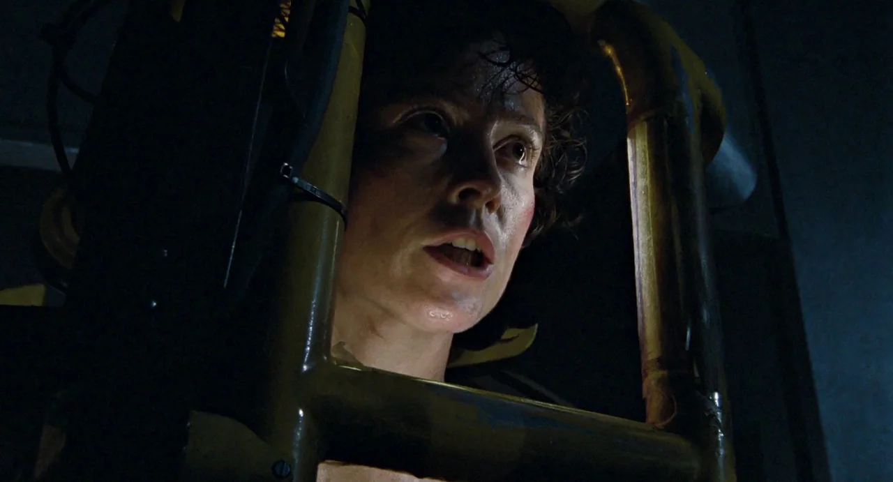Aliens (1986) movie still: closeup — A dark, cramped industrial environment featuring thick yellow mechanical pipes and framework. A woman with…