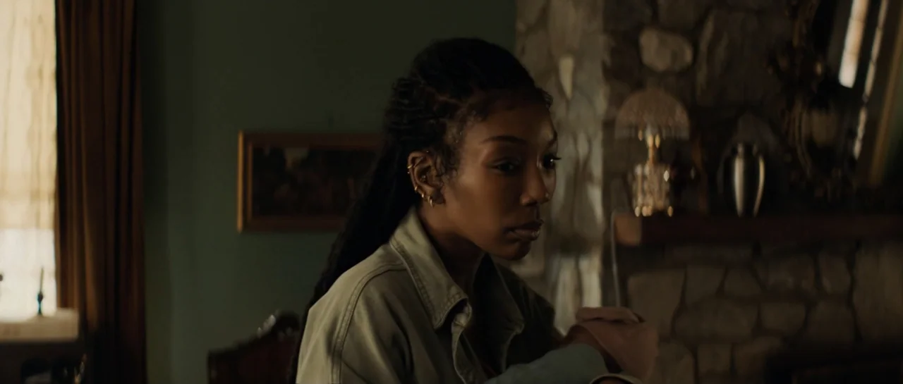 The Front Room (2024) movie still: medium — A dimly lit room features a stone fireplace and dark green walls. A young woman sits near the window and…