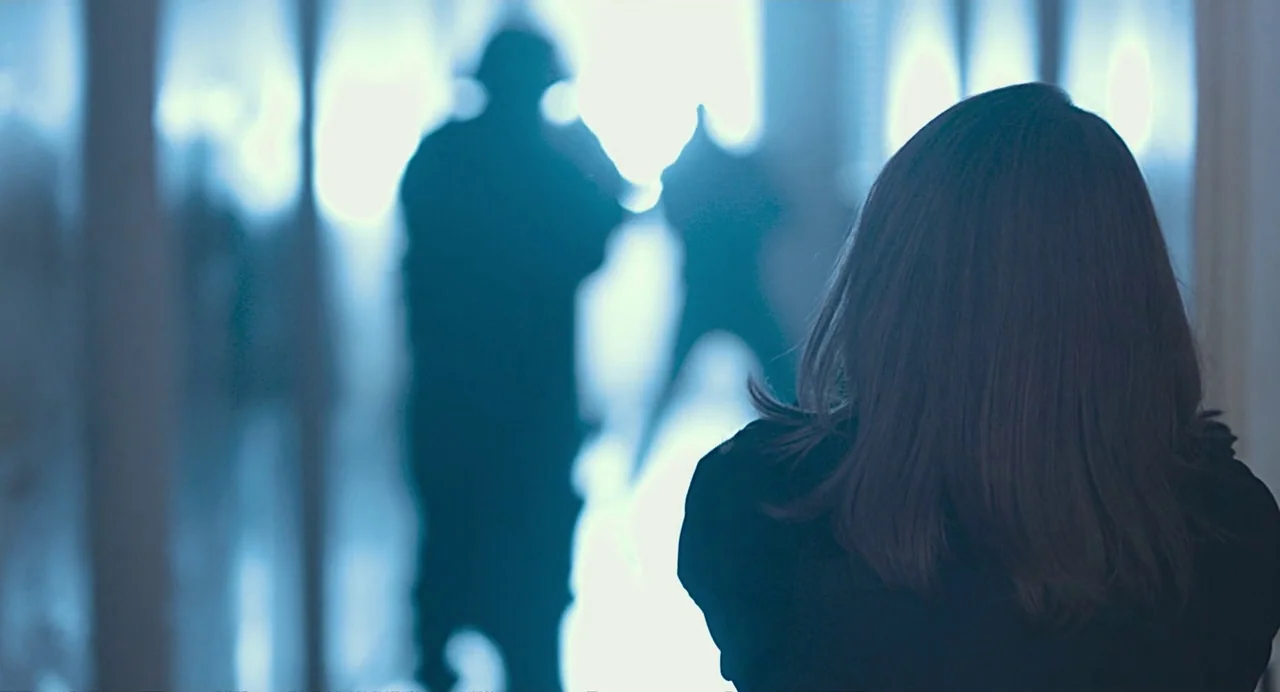 Non-Fiction (2018) movie still: medium — A dimly lit indoor hallway with a bright, overexposed light source at the end. A woman watches as two…