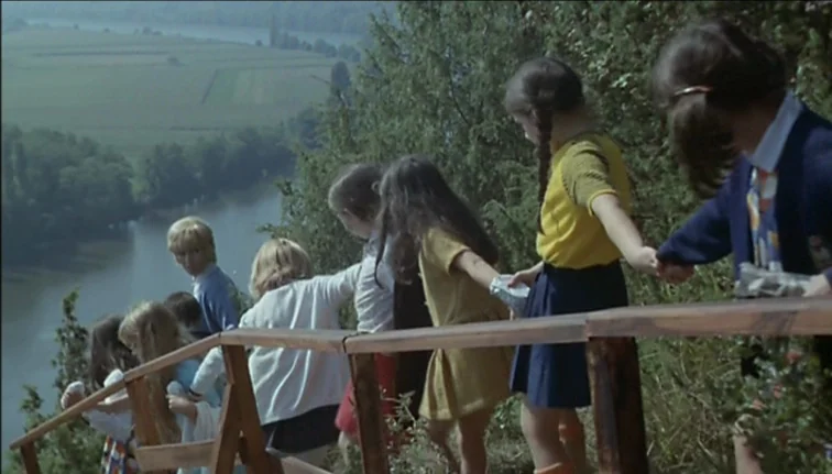 Le Boucher (1970) movie still: wide — A group of children walk along a wooden-railed path on a high river bluff. A winding river cuts through a…