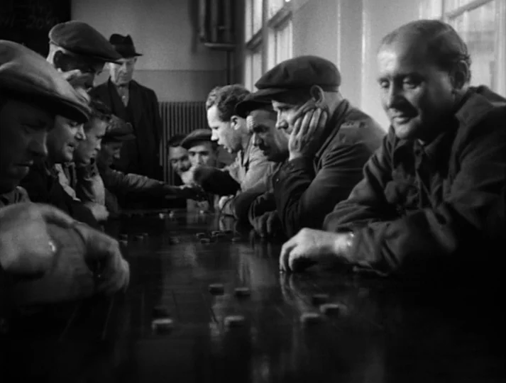 Port of Call (1948) movie still: medium — A group of men in flat caps sit at a long wooden game table. The men lean over the table and move small…