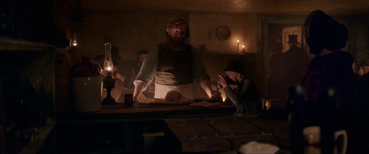 The Wonder (2022) movie still: medium — A dimly lit 19th-century establishment with dark wooden furniture and low ceilings. A bearded man works…
